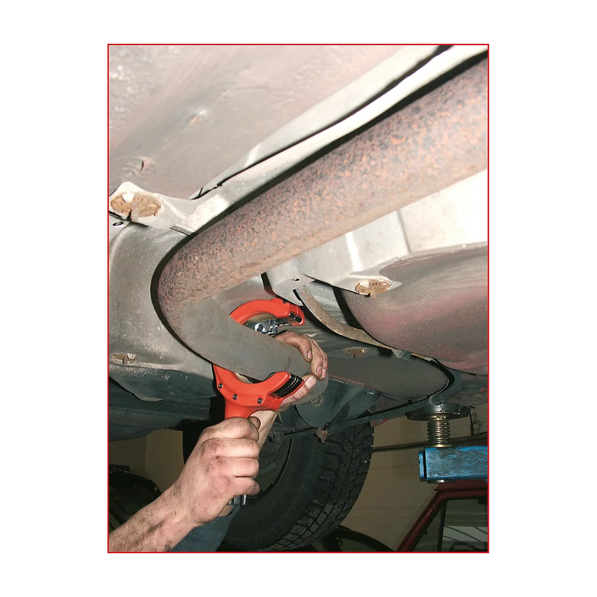 Ein Mechaniker schneidet ein Auspuffrohr mit einem roten Werkzeug unter einem Auto ab.